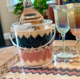 A decorative rectangular tray with a leopard print border and a chevron patterned center, holding a clear glass bottle in a clear glass bucket with a knit wrap, plus two iridescent fluted glasses, on a wooden table.